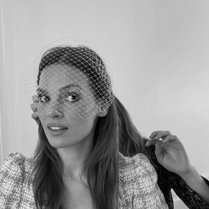 May include: A woman with long brown hair is wearing a white veil over her face. She is wearing a white and black tweed jacket.