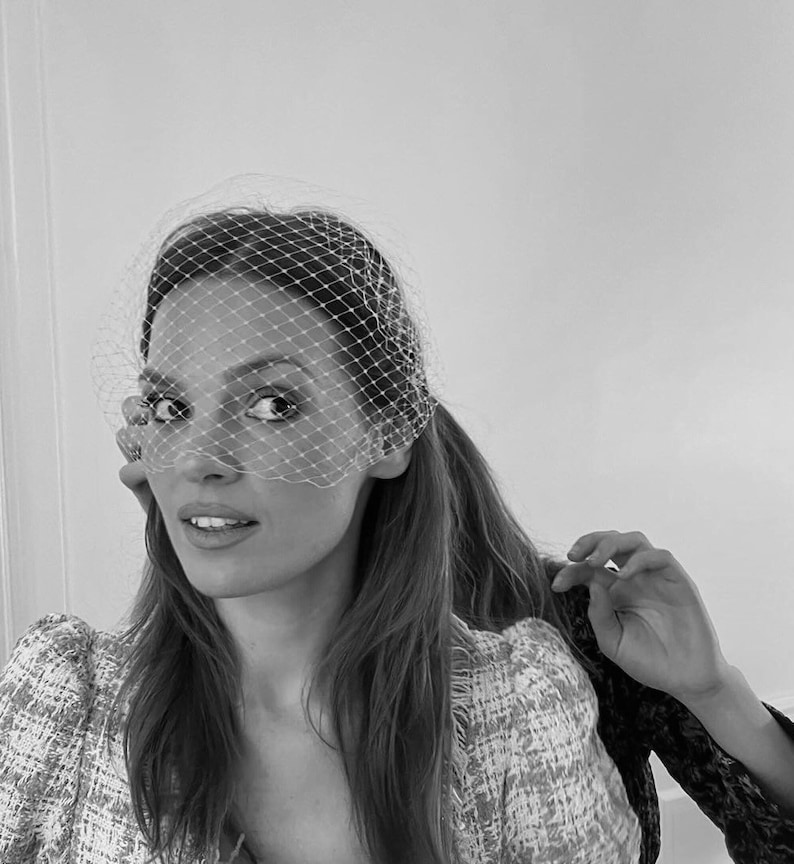 May include: A woman with long brown hair is wearing a white veil over her face. She is wearing a white and black tweed jacket.