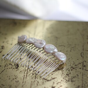May include: A silver hair comb with five white pearl-like embellishments. The comb is lying on a gold surface.