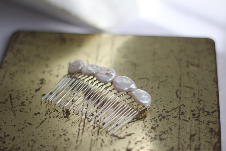May include: A silver hair comb with five white pearl-like embellishments. The comb is lying on a gold surface.