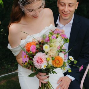 May include: A vibrant floral bouquet featuring pink and orange dahlias, white cosmos, and dark blackberries, held by a person in a white off-the-shoulder dress with a white birdcage veil, accompanied by a person in a dark suit and white shirt.
