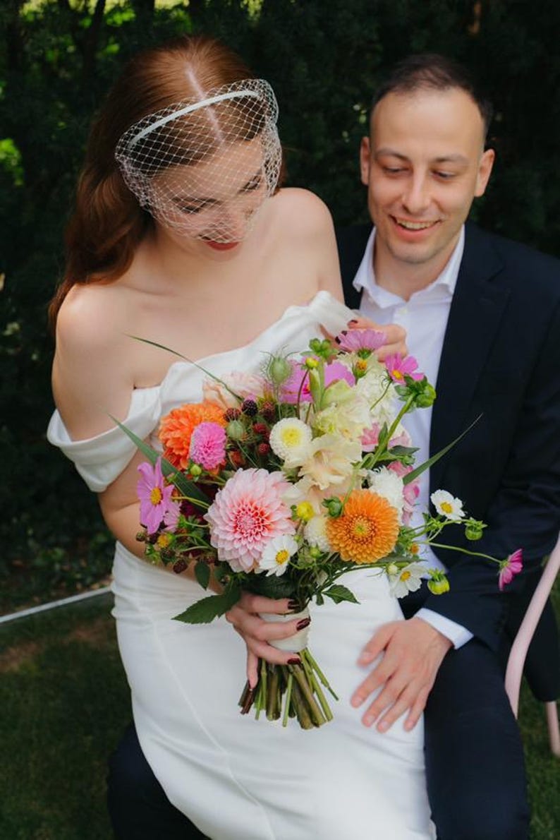 May include: A vibrant floral bouquet featuring pink and orange dahlias, white cosmos, and dark blackberries, held by a person in a white off-the-shoulder dress with a white birdcage veil, accompanied by a person in a dark suit and white shirt.
