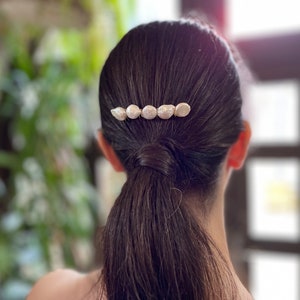 May include: A woman with long dark brown hair wearing a hair clip with five pearl-like white beads.