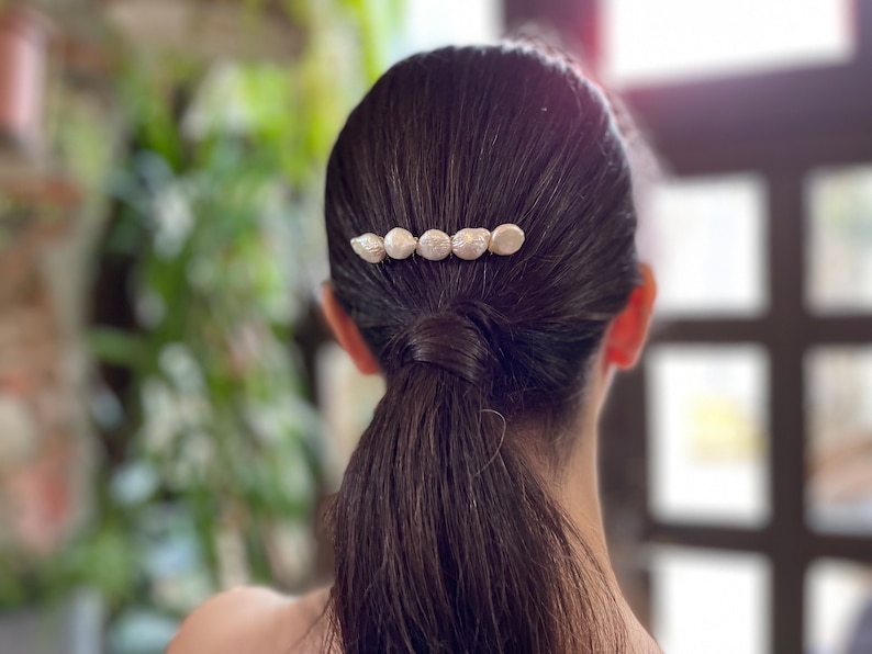 May include: A woman with long dark brown hair wearing a hair clip with five pearl-like white beads.