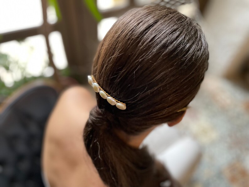 May include: A gold hair clip with a row of white oval pearls. The clip is in a woman's brown hair.