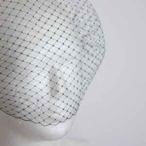 May include: A black birdcage veil made of fine netting. The veil is draped over a white mannequin head.