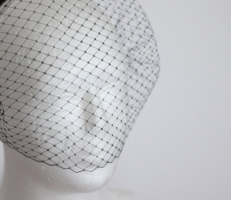 May include: A black birdcage veil made of fine netting. The veil is draped over a white mannequin head.