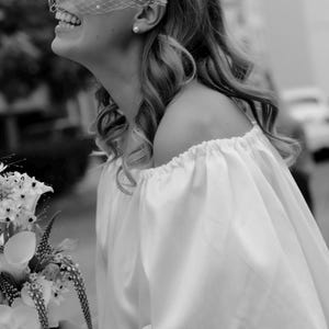 Op de afbeelding: Een witte off-shoulder jurk met geplooide mouwen en een bijpassende witte haarband met een birdcage-sluier versierd met kleine witte parels. Het geheel wordt gecombineerd met een boeket witte bloemen, waaronder calla lelies en orchidee&euml;n.