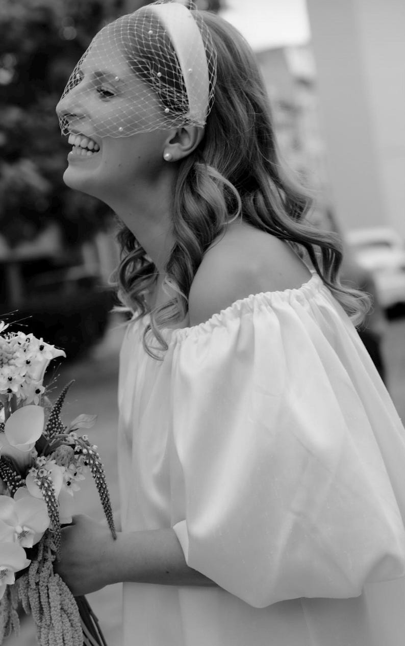 Op de afbeelding: Een witte off-shoulder jurk met geplooide mouwen en een bijpassende witte haarband met een birdcage-sluier versierd met kleine witte parels. Het geheel wordt gecombineerd met een boeket witte bloemen, waaronder calla lelies en orchidee&euml;n.