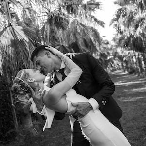 May include: A black and white photograph of two individuals in formal attire. One wears a sleeveless white gown with a row of buttons along the side and a birdcage veil. The other wears a dark suit with a tie and a decorative pin on the cuff.