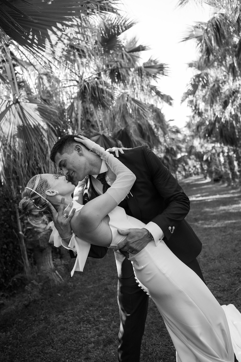 May include: A black and white photograph of two individuals in formal attire. One wears a sleeveless white gown with a row of buttons along the side and a birdcage veil. The other wears a dark suit with a tie and a decorative pin on the cuff.