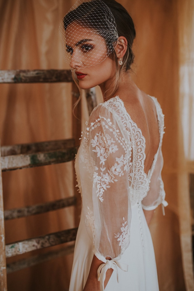 May include: A bride wearing a white lace wedding dress with a low back and sheer sleeves. The dress has a delicate floral pattern and the sleeves are tied at the wrist. The bride is wearing a veil and has her hair pulled back.