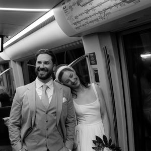 Op de afbeelding: Een zwart-witfoto van twee personen in formele kleding in een metrostel. De een draagt een driedelig pak met stropdas, de ander een mouwloze jurk met een birdcage-sluier en houdt een boeket bloemen vast bij een kaart van het metronetwerk.