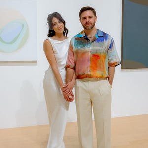 May include: A couple poses in front of abstract paintings in a gallery. The woman is wearing a white dress and a veil. The man is wearing a colorful patterned shirt and beige pants.