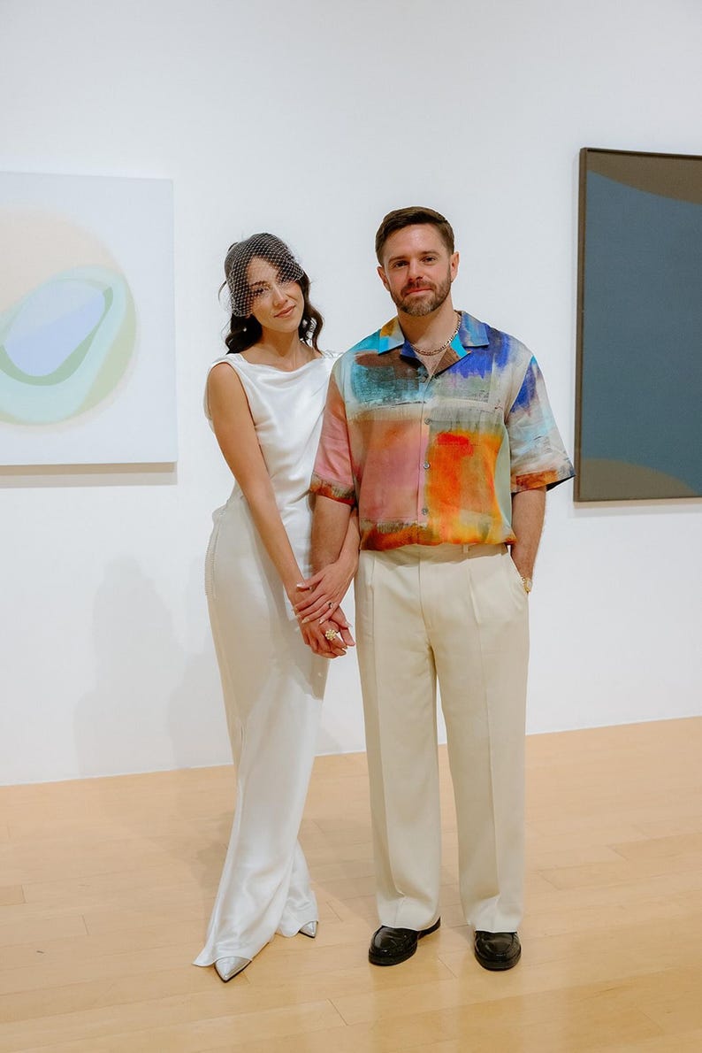 May include: A couple poses in front of abstract paintings in a gallery. The woman is wearing a white dress and a veil. The man is wearing a colorful patterned shirt and beige pants.