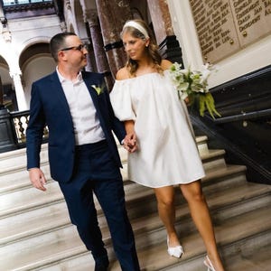Op de afbeelding: Een marineblauw pak met een wit overhemd en zwarte schoenen gecombineerd met een off-shoulder witte minijurk, witte slingback-hakken en een witte haarband met een birdcage-sluier, terwijl een boeket witte bloemen wordt vastgehouden.