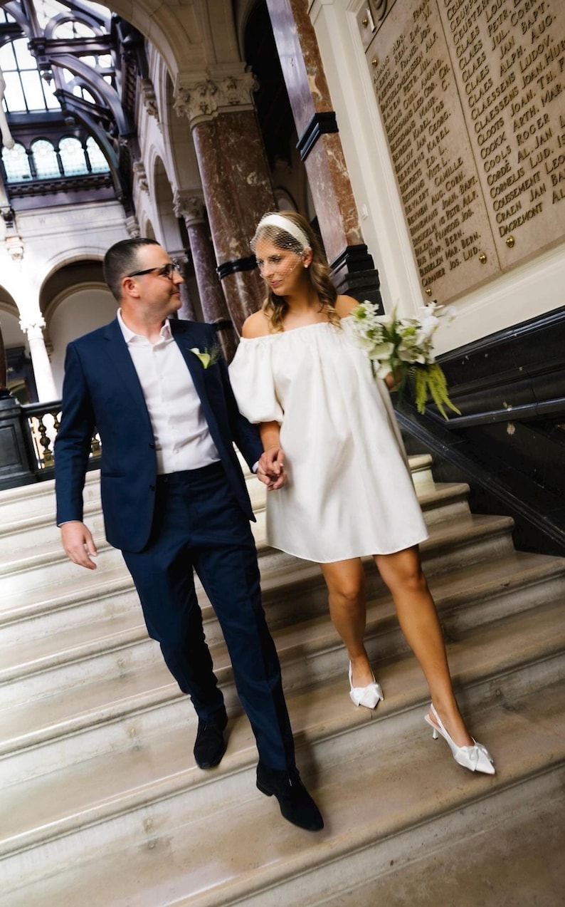 Op de afbeelding: Een marineblauw pak met een wit overhemd en zwarte schoenen gecombineerd met een off-shoulder witte minijurk, witte slingback-hakken en een witte haarband met een birdcage-sluier, terwijl een boeket witte bloemen wordt vastgehouden.