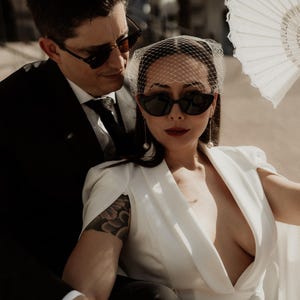 May include: A couple wearing sunglasses and a white dress with a veil. The woman has a tattoo on her arm. The man is wearing a black suit and tie.