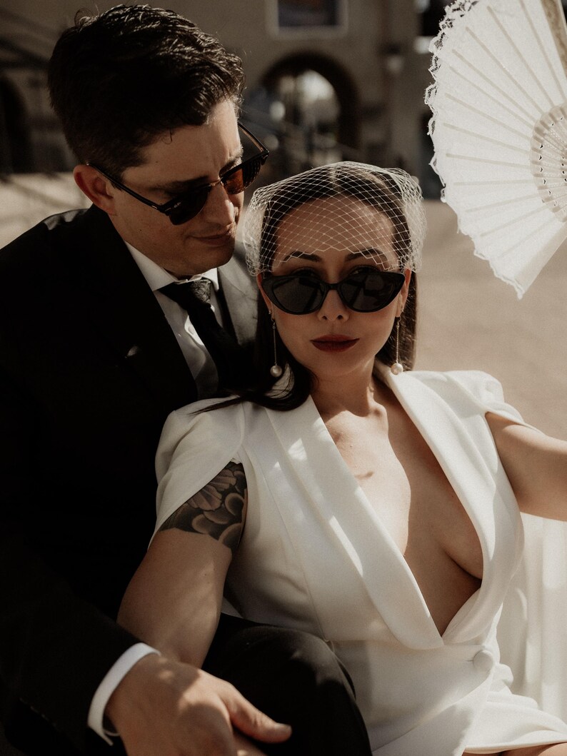 May include: A couple wearing sunglasses and a white dress with a veil. The woman has a tattoo on her arm. The man is wearing a black suit and tie.