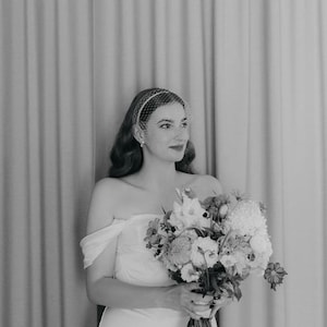 May include: A black and white photograph of a strapless gown with off-the-shoulder sleeves and a birdcage veil. The garment is paired with a large, textured floral bouquet featuring various blossoms and stems held in the center of the frame.