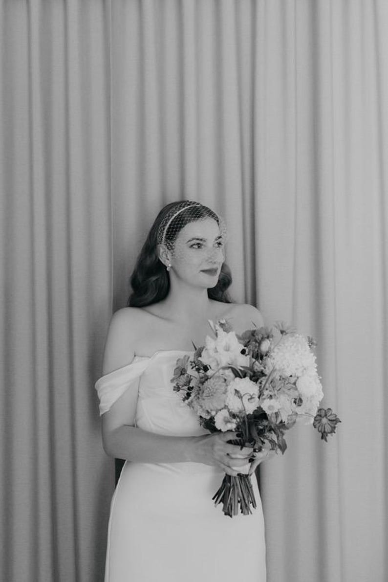 May include: A black and white photograph of a strapless gown with off-the-shoulder sleeves and a birdcage veil. The garment is paired with a large, textured floral bouquet featuring various blossoms and stems held in the center of the frame.