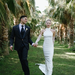 May include: A dark navy blue suit with a white shirt, dark tie, and boutonniere paired with a floor-length white halter-neck gown featuring sheer long sleeves and a train, worn by two individuals walking together on a grassy path.