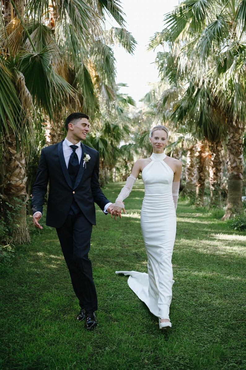 May include: A dark navy blue suit with a white shirt, dark tie, and boutonniere paired with a floor-length white halter-neck gown featuring sheer long sleeves and a train, worn by two individuals walking together on a grassy path.