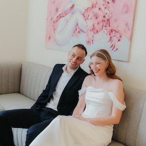 May include: A white off-the-shoulder wedding dress with a structured bodice and a floor-length skirt, paired with a white birdcage veil. The ensemble is worn alongside a black suit jacket, a white button-down shirt, and black trousers.