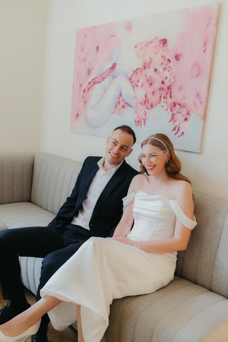 May include: A white off-the-shoulder wedding dress with a structured bodice and a floor-length skirt, paired with a white birdcage veil. The ensemble is worn alongside a black suit jacket, a white button-down shirt, and black trousers.