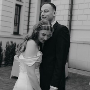 May include: A black and white photograph of two individuals standing together. One wears a white off-the-shoulder gown with a birdcage veil over their face, while the other wears a dark suit jacket and shirt. They are holding hands closely.