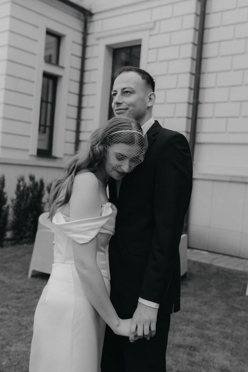 May include: A black and white photograph of two individuals standing together. One wears a white off-the-shoulder gown with a birdcage veil over their face, while the other wears a dark suit jacket and shirt. They are holding hands closely.