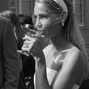 Op de afbeelding: Een witte haarband met een birdcage-sluier versierd met kleine witte parels. De sluier bedekt het bovenste deel van het gezicht en loopt vanaf de haarband naar beneden. Het accessoire wordt gedragen met een wit kledingstuk zonder schouderbandjes.