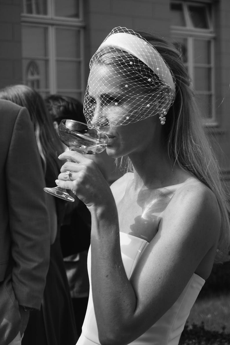 Op de afbeelding: Een witte haarband met een birdcage-sluier versierd met kleine witte parels. De sluier bedekt het bovenste deel van het gezicht en loopt vanaf de haarband naar beneden. Het accessoire wordt gedragen met een wit kledingstuk zonder schouderbandjes.