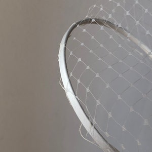 May include: A white headband featuring a delicate, fine-mesh netting overlay. The netting is constructed with a repeating diamond-shaped pattern, and the headband itself is covered in a smooth, solid white fabric material.