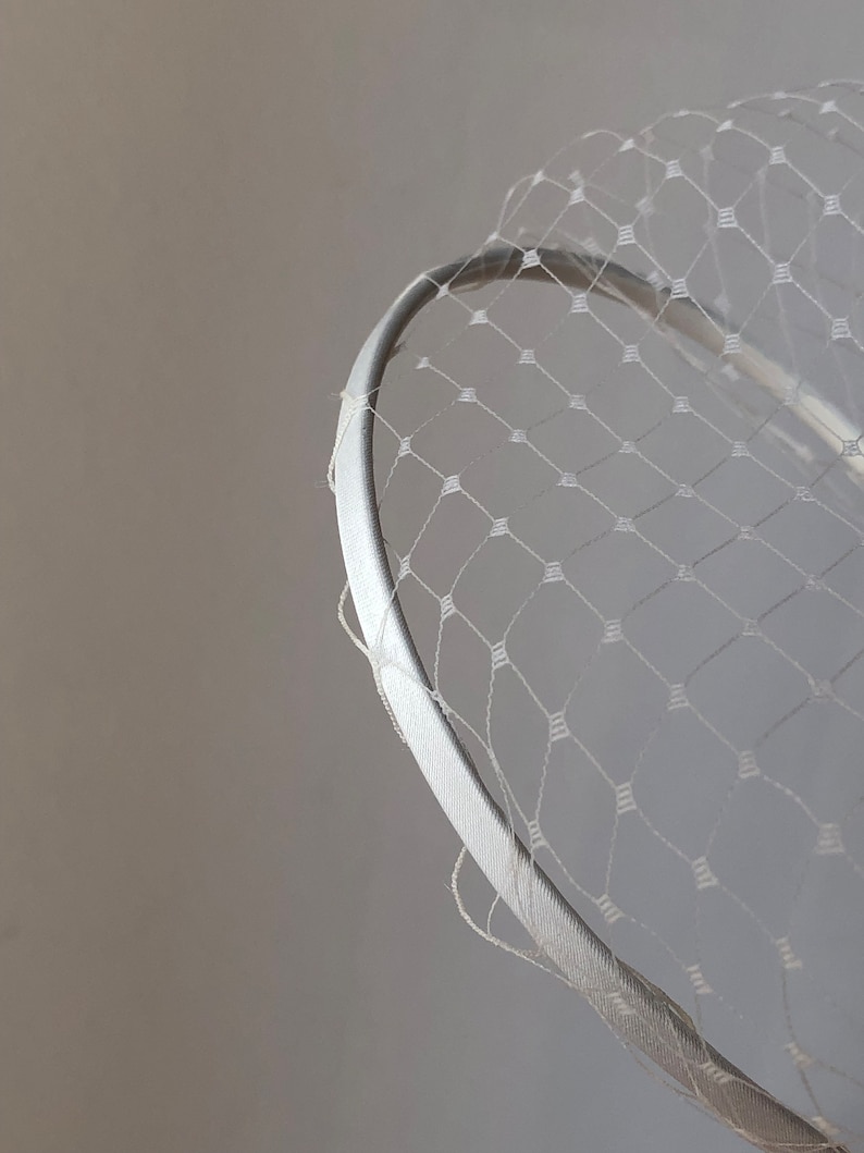 May include: A white headband featuring a delicate, fine-mesh netting overlay. The netting is constructed with a repeating diamond-shaped pattern, and the headband itself is covered in a smooth, solid white fabric material.