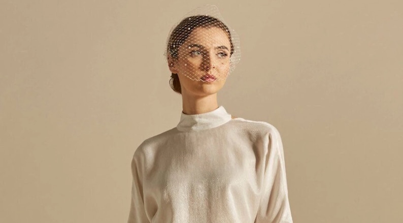 May include: A white high-neck long-sleeve top with a subtle shimmer paired with a white birdcage veil featuring small evenly spaced pearls. The veil is positioned over the upper face, while the top has a structured collar and long sleeves.