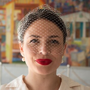 May include: A woman wearing a white veil with a delicate net pattern over her face. She is wearing a white shirt and has red lipstick on.