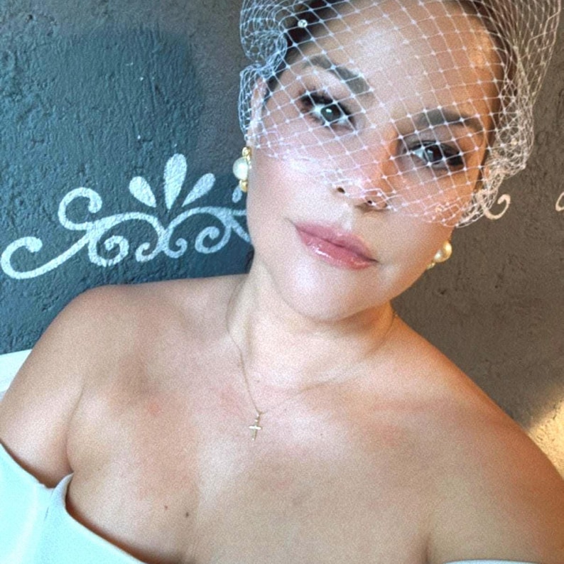 May include: A white birdcage veil featuring a fine mesh net pattern with small embellishments, paired with round cream-colored stud earrings and a delicate gold chain necklace with a small cross pendant, worn over a plain white off-the-shoulder garment.