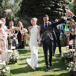 May include: A couple walking through a shower of confetti after a wedding ceremony. The person on the left wears a white floor-length halter-neck gown and holds a bouquet of pink and white flowers. The person on the right wears a dark suit.
