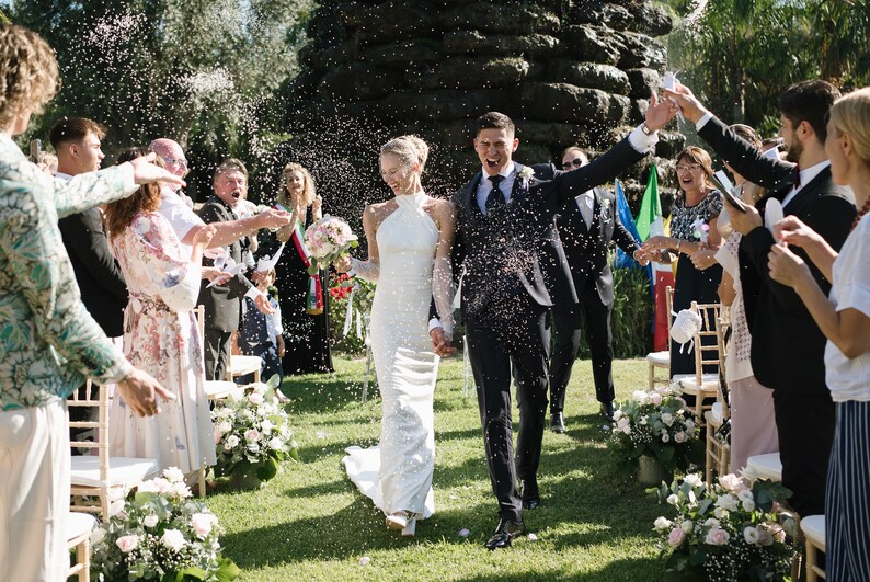 May include: A couple walking through a shower of confetti after a wedding ceremony. The person on the left wears a white floor-length halter-neck gown and holds a bouquet of pink and white flowers. The person on the right wears a dark suit.