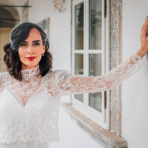 May include: A woman wearing a white lace dress with a veil and red lipstick. She is standing in front of a white wall with a window behind her.