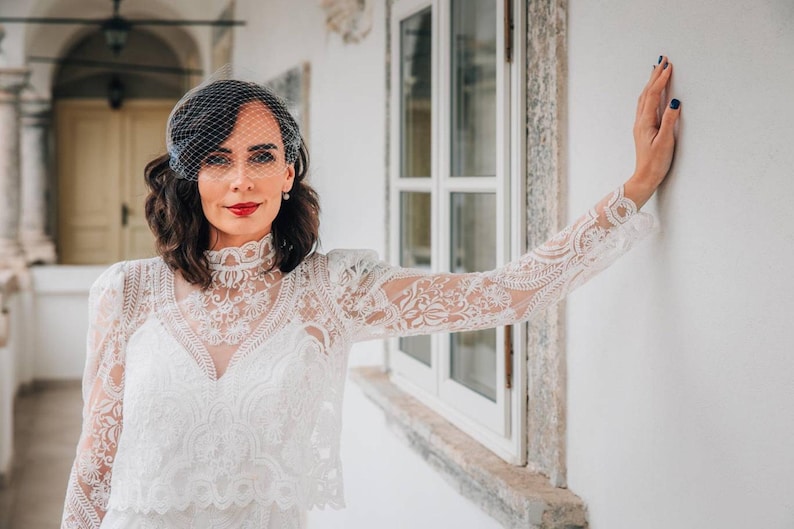 May include: A woman wearing a white lace dress with a veil and red lipstick. She is standing in front of a white wall with a window behind her.