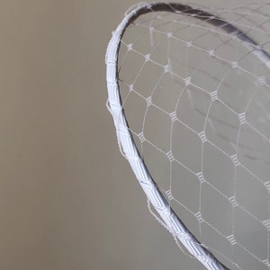 May include: A white birdcage veil featuring a fine, diamond-patterned netting attached to a curved, ribbon-wrapped headband. The delicate mesh fabric creates a structured, semi-transparent layer that arches outward from the smooth, fabric-covered frame.