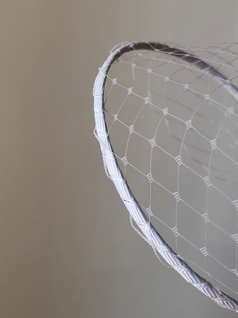 May include: A white birdcage veil featuring a fine, diamond-patterned netting attached to a curved, ribbon-wrapped headband. The delicate mesh fabric creates a structured, semi-transparent layer that arches outward from the smooth, fabric-covered frame.