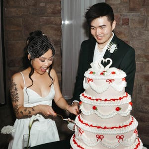 May include: A bride and groom cutting a white wedding cake decorated with red and white frosting, swans, and bows. The cake has four tiers and is on a white table.