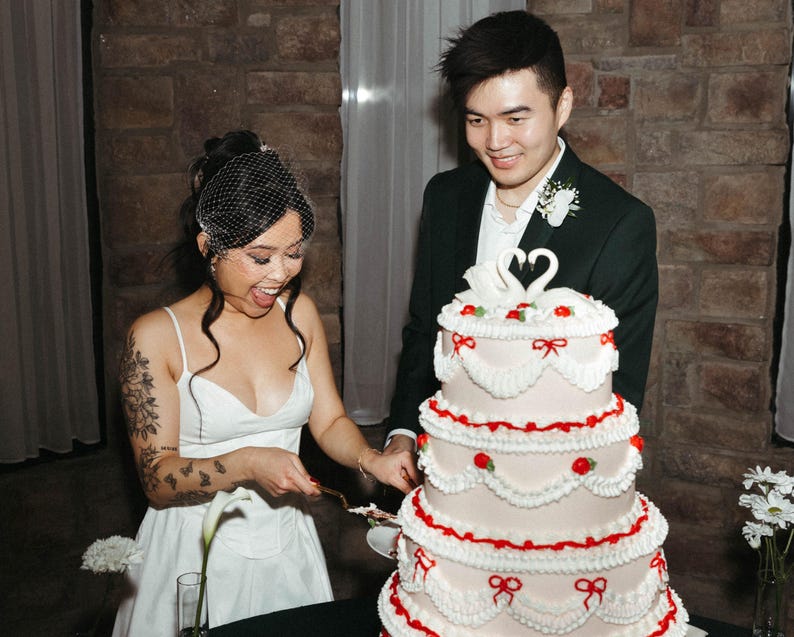 May include: A bride and groom cutting a white wedding cake decorated with red and white frosting, swans, and bows. The cake has four tiers and is on a white table.