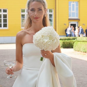 Op de afbeelding: Een strapless witte jurk gecombineerd met een bijpassende witte hoofdband met een delicate birdcage-sluier. Het ensemble bevat een wit boeket hortensia's, een wit jasje over &eacute;&eacute;n arm en een helder glazen coupe voor een drankje.
