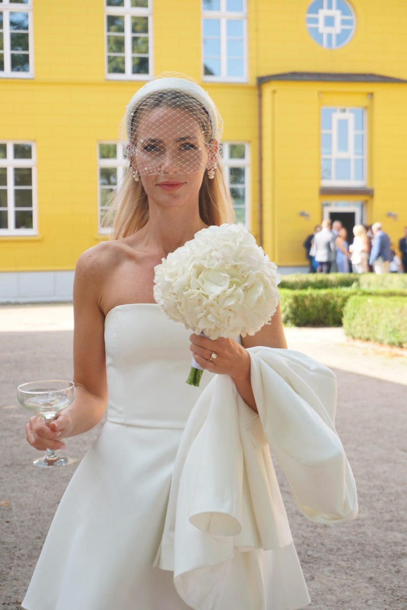 Op de afbeelding: Een strapless witte jurk gecombineerd met een bijpassende witte hoofdband met een delicate birdcage-sluier. Het ensemble bevat een wit boeket hortensia's, een wit jasje over &eacute;&eacute;n arm en een helder glazen coupe voor een drankje.