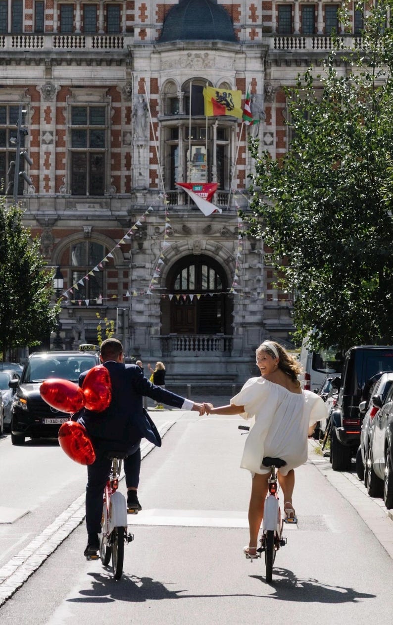 Op de afbeelding: Twee personen fietsen op een stadsstraat terwijl ze elkaars hand vasthouden. E&eacute;n persoon draagt een donkerblauw pak en heeft drie rode hartvormige ballonnen bij zich. De andere persoon draagt een korte witte jurk en een witte haarband. Een groot stenen gebouw staat op de achtergrond.