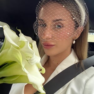 Op de afbeelding: Een witte haarband met een fijne birdcage-sluier versierd met kleine witte parels. Het accessoire wordt gecombineerd met een witte blazer en een boeket witte calla-lelies, wat zorgt voor een samenhangende uitstraling met parelhangers.